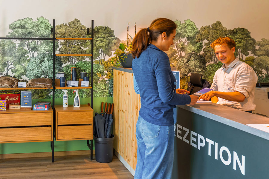 Freundlicher Check-in an der Rezeption mit Waldtapete, Gästin beim Einchecken