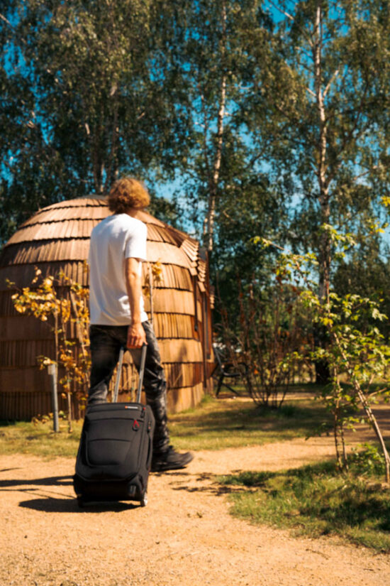 Gast reist mit Koffer an und läuft zum Spiegelhaus im Wald mit runder Holzschindel-Fassade