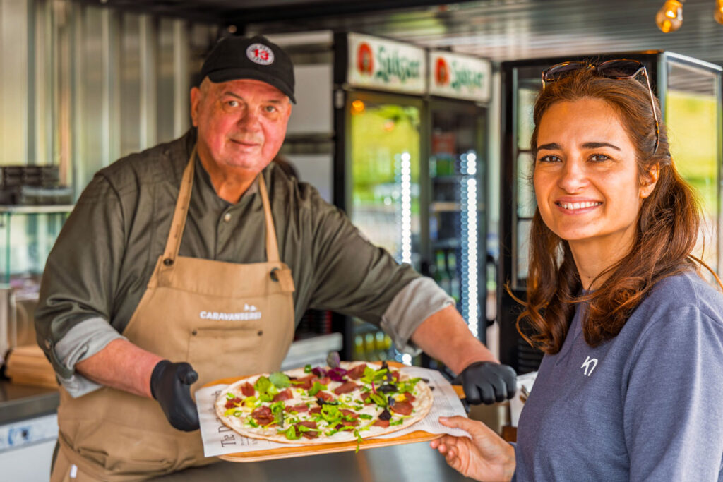 Mitarbeiter reicht einer Gästin frischen Flammkuchen im Sommergarten der CARAVANSEREI